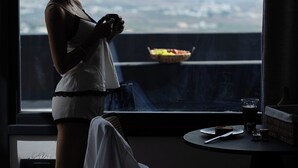 Una habitación con un ventanal grande que da a un paisaje urbano, una mesita con un vaso de agua y una silla.