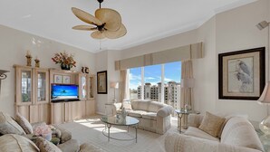 Ein geräumiges Wohnzimmer mit großem Flachbildfernseher, bequemen Sitzgelegenheiten, Deckenventilator und Blick auf die Stadt durch große Fenster.