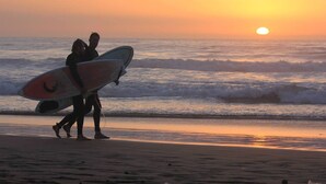 Deux personnes se promènent sur une plage, portant des planches de surf, et se dirigent vers l’océan au coucher du soleil.