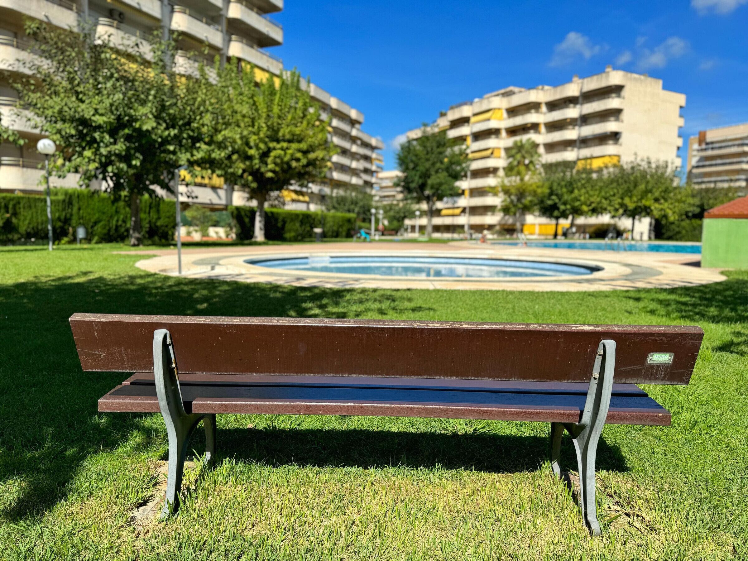 A park bench in a grassy area with a circular fountain and multi-story buildings in the background.