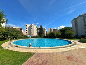 Ein großer, runder Swimmingpool mit klar blauem Wasser, umgeben von einem gepflasterten Weg und üppigem Grün. Im Hintergrund sind mehrstöckige Wohngebäude mit Balkonen zu sehen.