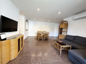 Ein Wohnzimmer mit Flachbildfernseher, einer Couch, einem Couchtisch, einem Esstisch mit Stühlen und einem Korbschrank.