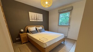 A bedroom with a wooden bed, a nightstand, a wall-mounted lamp, a window with a view of trees, and a painting of sailboats. - Two-room apartment in Sète - Terrace, Parking, WiFi (Sète)