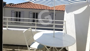 A balcony with a white table and chairs, a striped umbrella, and a building with a red-tiled roof. - Cozy studio downtown near the beach (La Tranche-sur-Mer)
