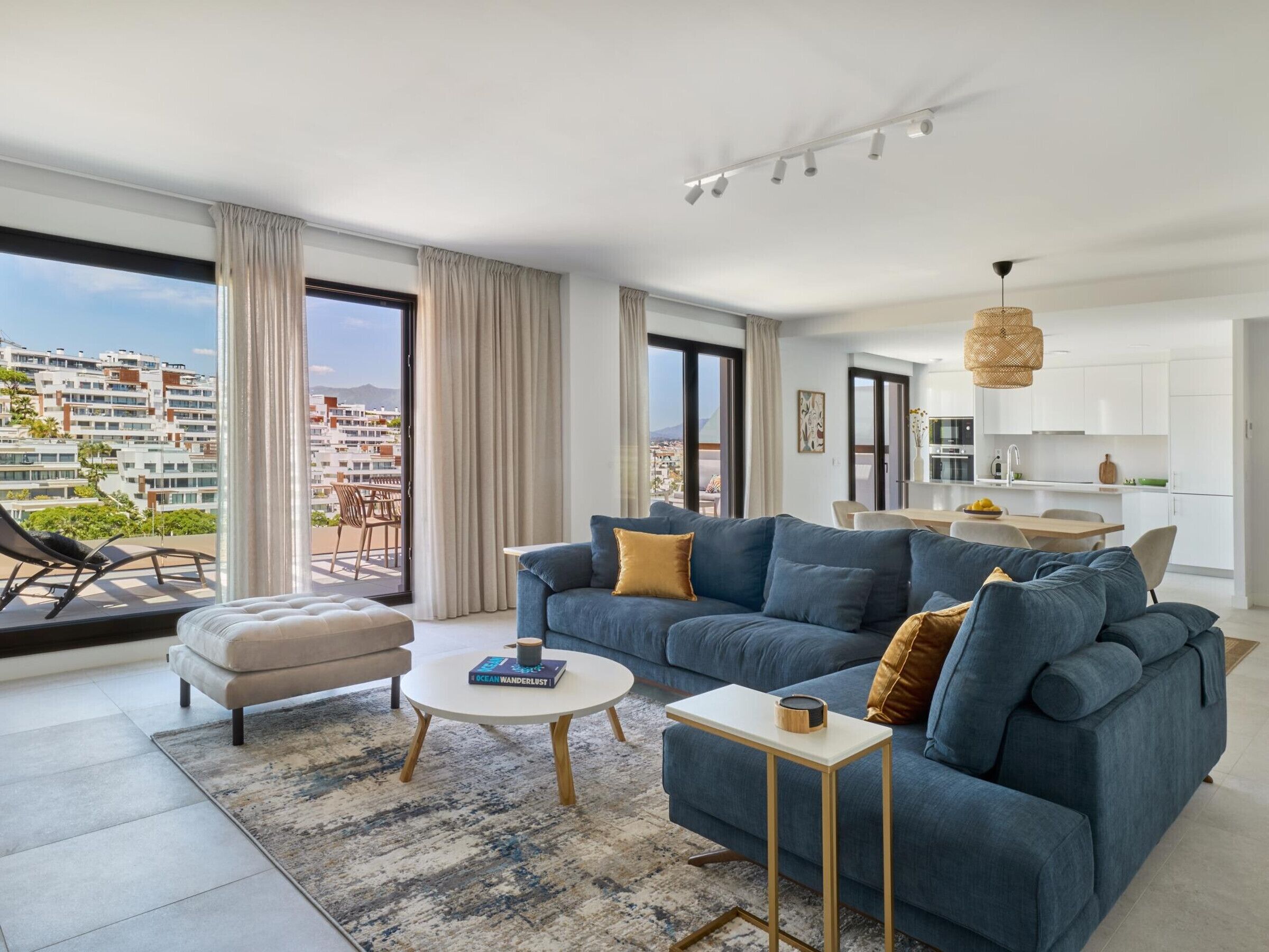 A modern living room with a sofa, ottoman, and coffee table, a balcony with a view of buildings, and a kitchen area.