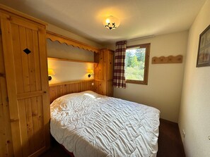A bedroom with a wooden headboard, a bed with a white quilt, a window with a plaid curtain, and a wall-mounted lamp. - Apartment for 4, ski-in/ski-out with balcony, parking included (Lanslebourg-Mont-Cenis)