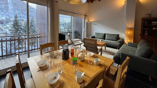 A dining area with a wooden table set for four, a sofa, and a view of a snowy landscape through large windows. - Spacious apartment for 8, close to slopes in Serre Chevalier (La Salle-les-Alpes)