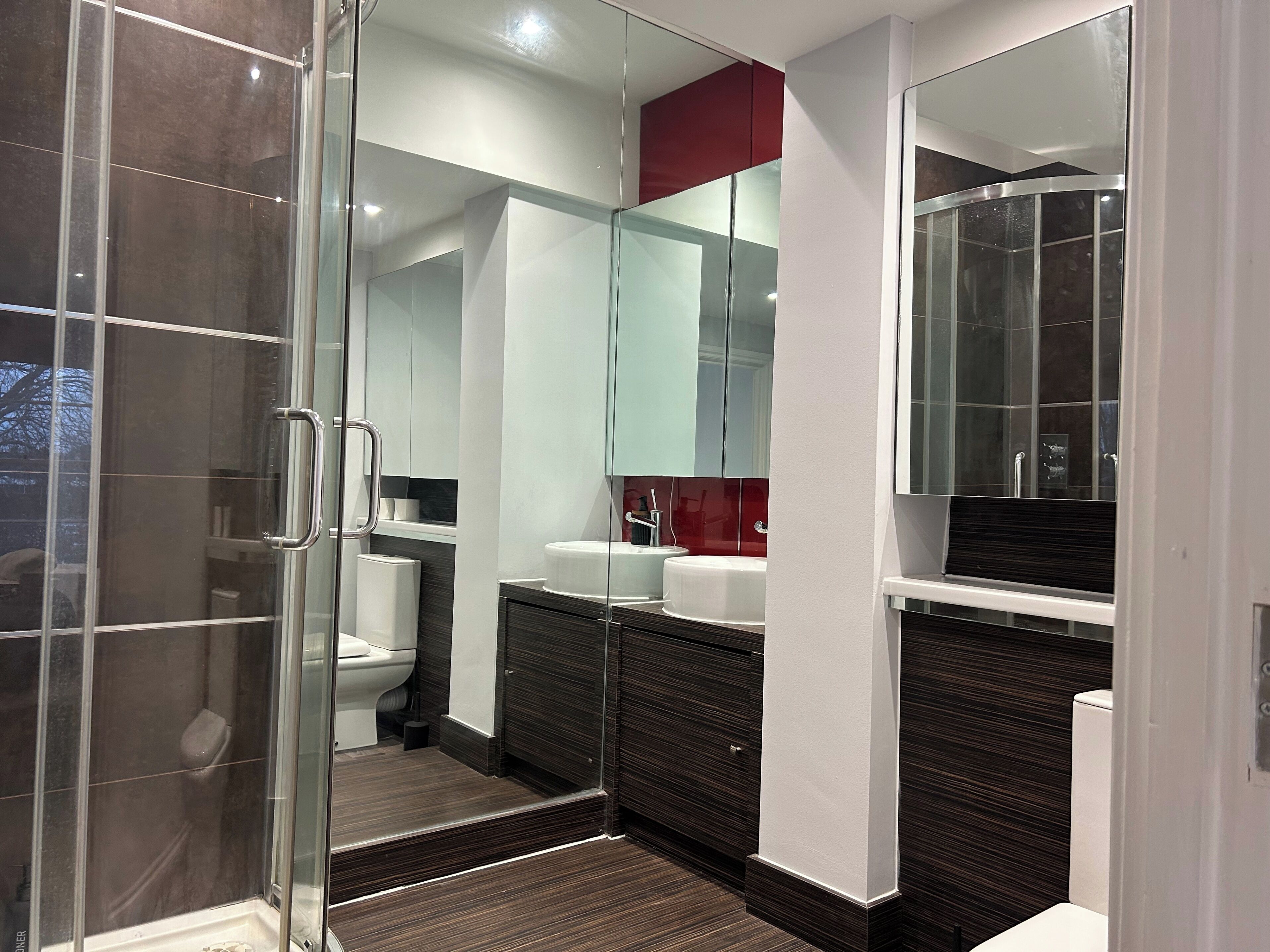 A modern bathroom with a glass shower enclosure, a white toilet, a vanity with a red accent, and a large mirror.