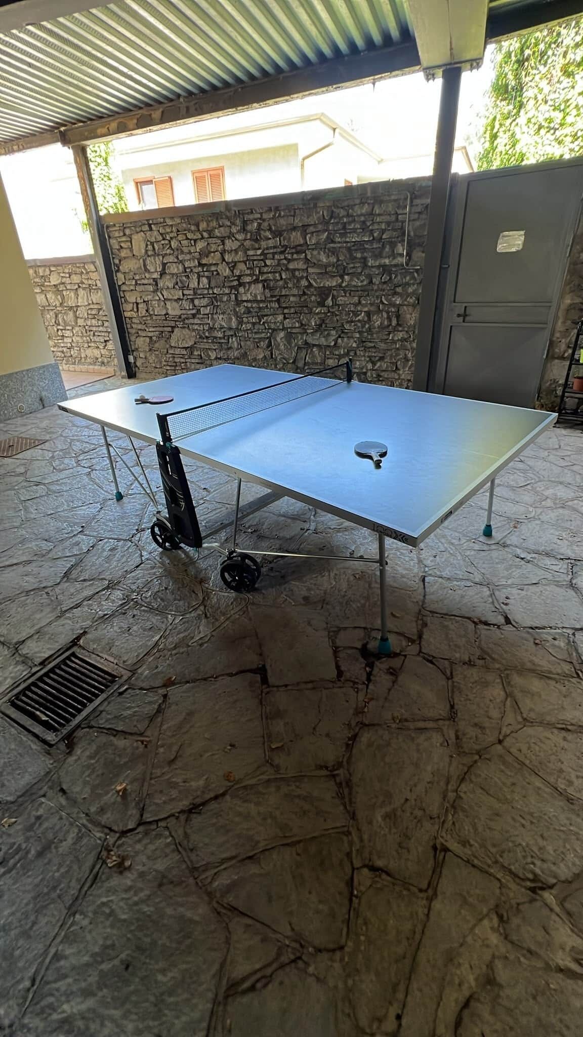 Ein Tischtennis-Tisch auf Rollen, auf einer Steinterrasse unter einem Metalldach platziert.