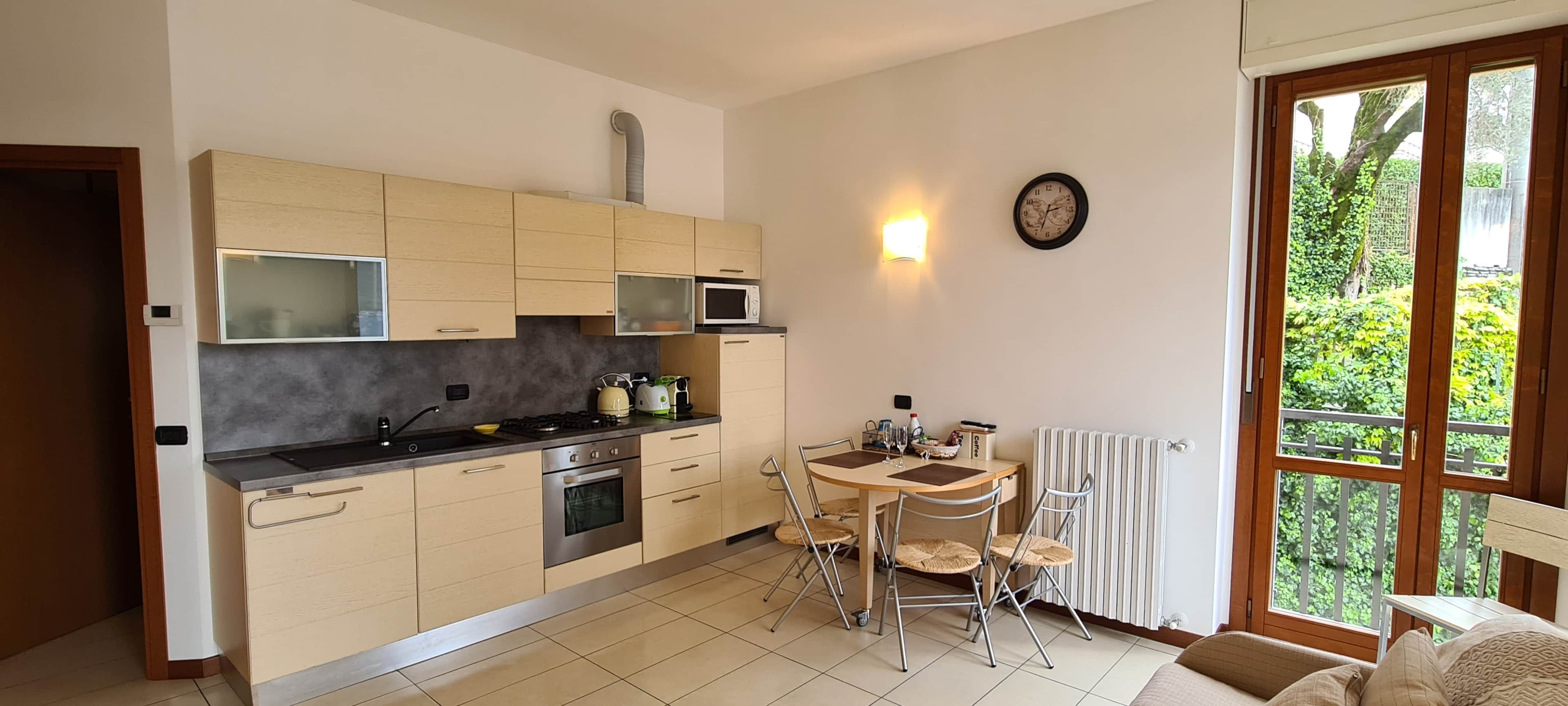 Une cuisine moderne avec un coin repas, une horloge au mur et une vue sur la verdure à l’extérieur.