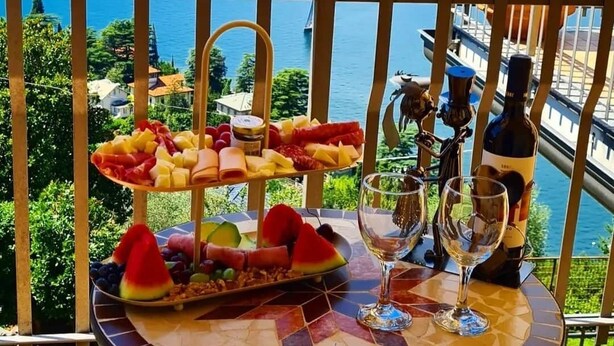 Ein Balkon mit Blick auf einen See und Berge, ein Tisch mit einer Weinflasche, Gläsern und einer Obstplatte.