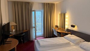 A hotel room with a bed, desk, chair, and a small table. There is a window with curtains, a lamp, and a wall-mounted light. - Roomie Hotel (Schriesheim)
