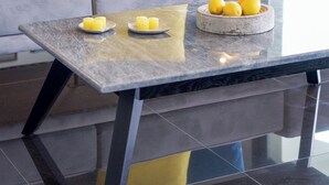 Una sala moderna con un sofá gris, cojines amarillos y azules, una mesa de centro de vidrio con un cuenco de limones y una planta en maceta al fondo.