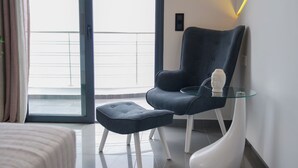 Una habitación de hotel moderna con un ventanal grande, un sillón azul oscuro, una mesa de centro de vidrio y una mesita de noche blanca de diseño escultórico.