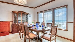 Een eetkamer met een grote tafel, zes stoelen, een kroonluchter, een houten kast en uitzicht op de oceaan door grote ramen.