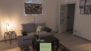 Un salon avec un canapé gris, une table basse, une projection d’une carte du monde sur le mur, une petite table d’appoint avec une lampe, et une porte donnant sur une autre pièce.