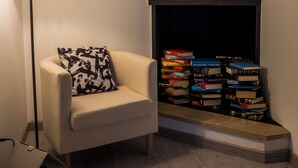 Un salon chaleureux avec un fauteuil blanc, une lampe sur pied, une cheminée surmontée d’une pile de livres et un tableau encadré accroché au mur.