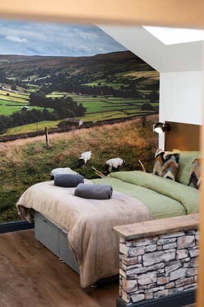 Une chambre avec une fresque représentant un paysage verdoyant, un lit avec des oreillers, un mur en pierre servant d’élément décoratif et une lampe de chevet.