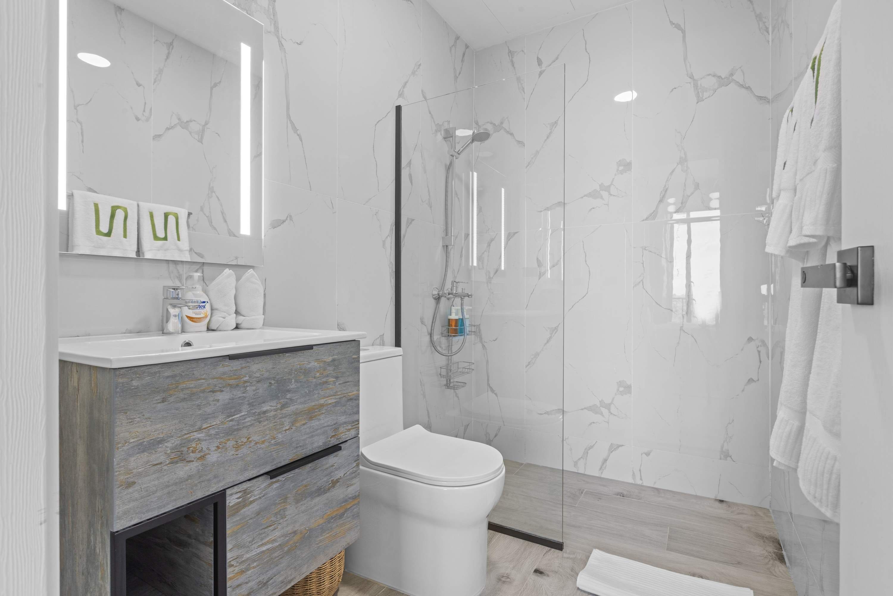 A modern bathroom with a marble wall, a wooden vanity, a white toilet, and a glass shower enclosure.