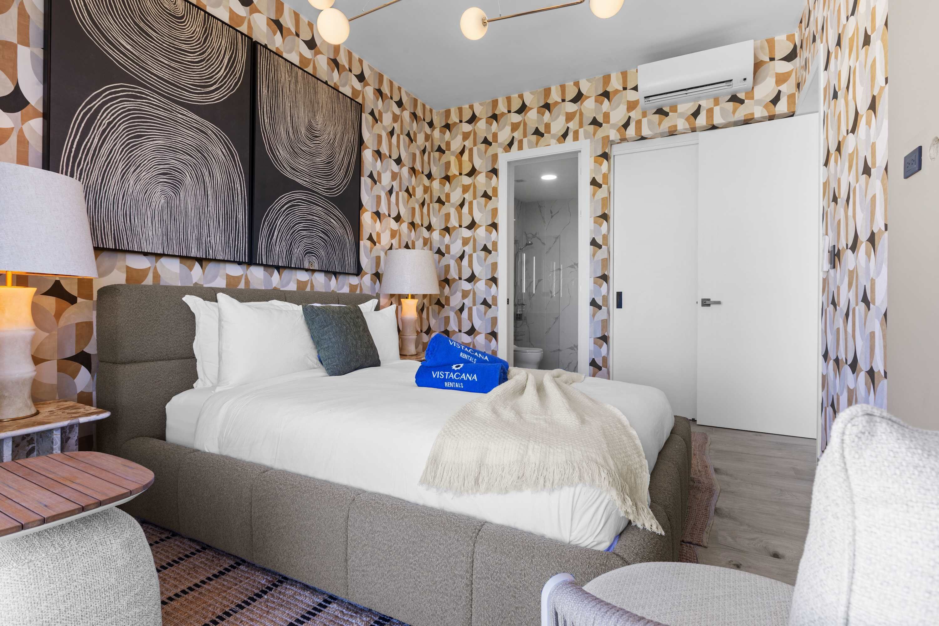 A modern hotel room with a large bed, two bedside lamps, a small table, and a chair. There are two blue 'Vistacana Rentals' bags on the bed.