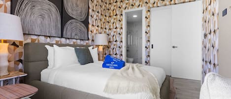 Una camera d'albergo moderna con un letto grande, due lampade ai comodini, un tavolino e una sedia. Ci sono due borse blu di 'Vistacana Rentals' sul letto.