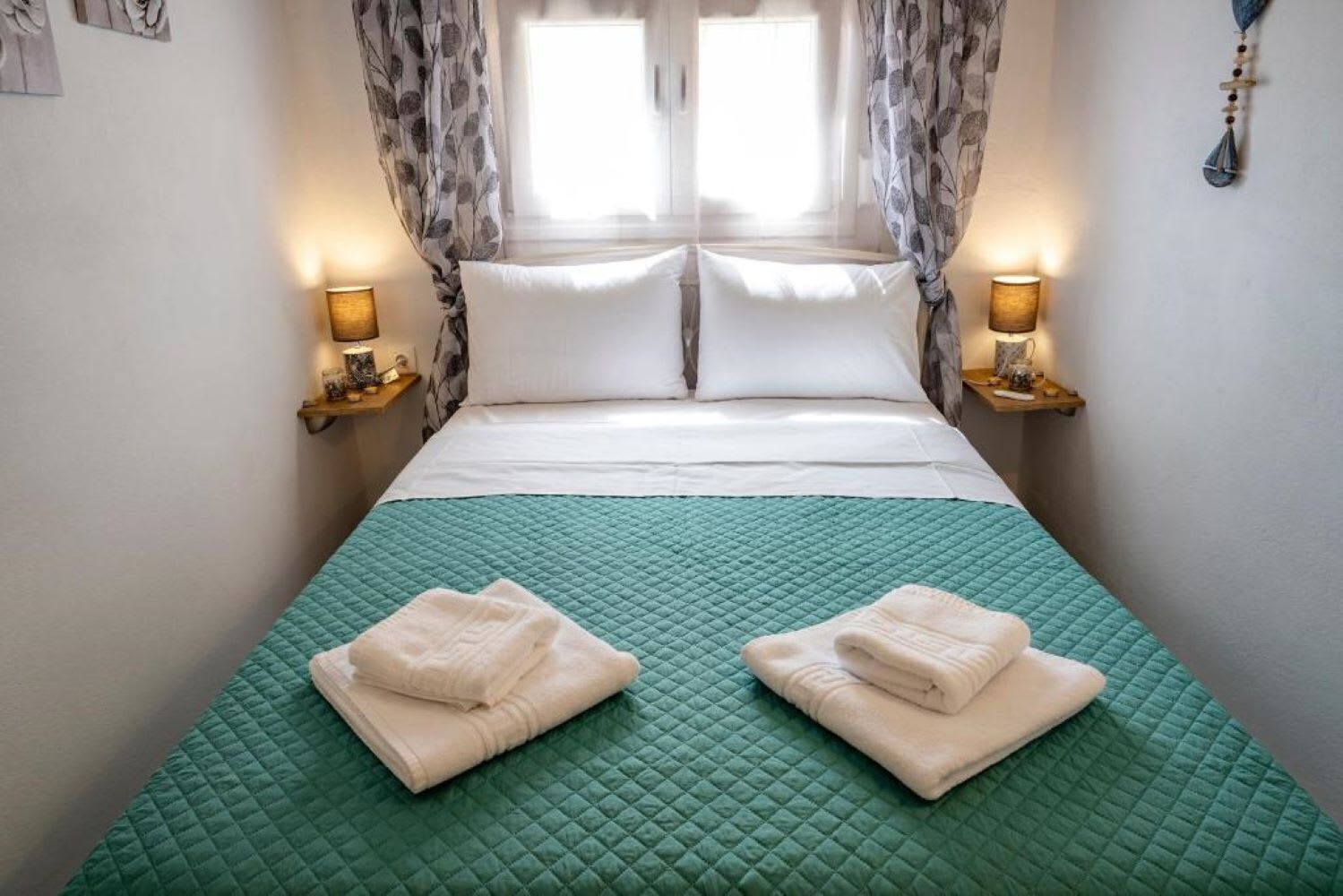 Una cama bien hecha con un edredón verde, dos toallas dobladas y lámparas de noche.
