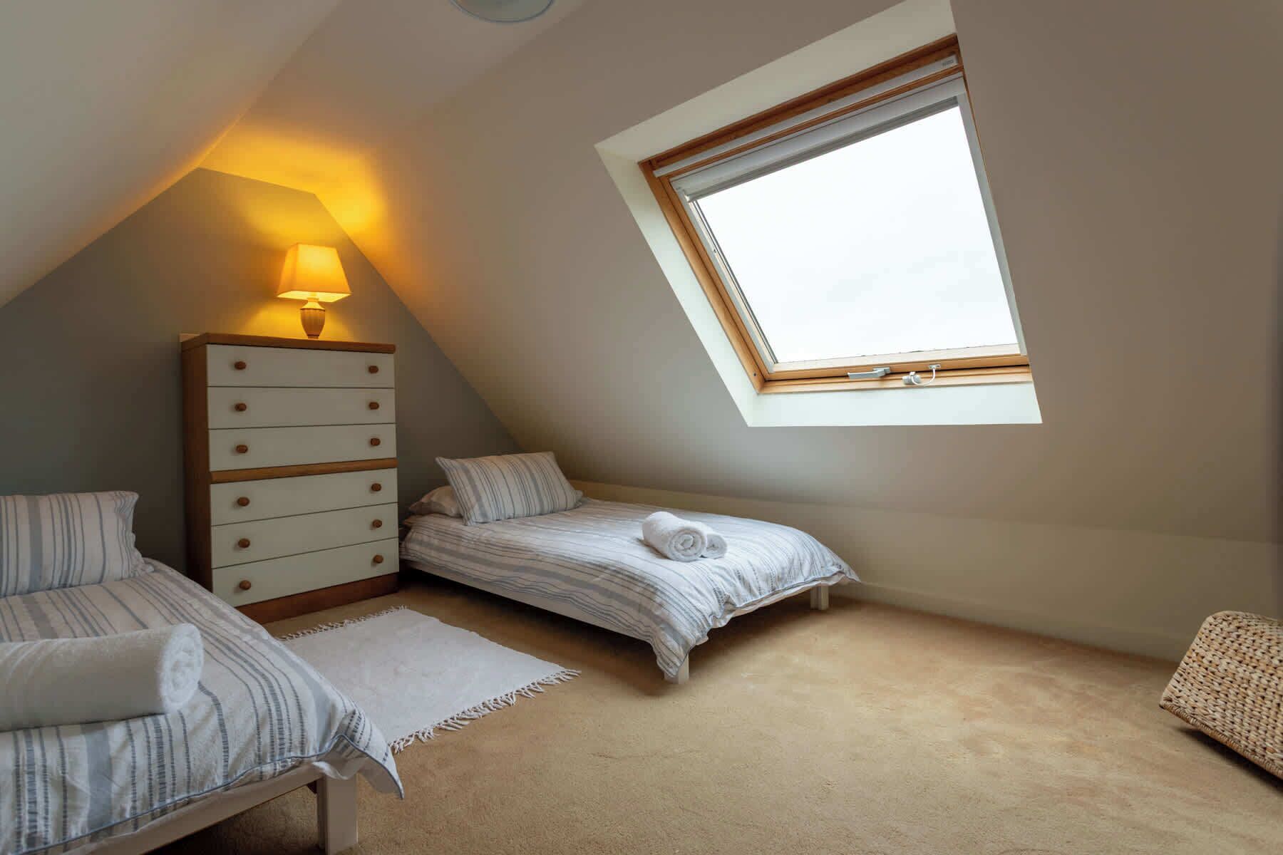 A small, cozy attic bedroom with a sloped ceiling, a skylight, a wooden dresser, two beds, and a small rug.