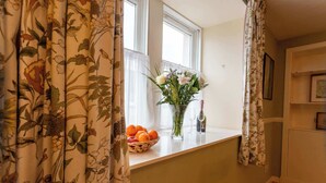 A room with a window, a vase with flowers, a bottle, and a basket of oranges on the windowsill. - New Cross Keys House - Cosy 4-bed Cottage in Kirk Yetholm (Kelso)