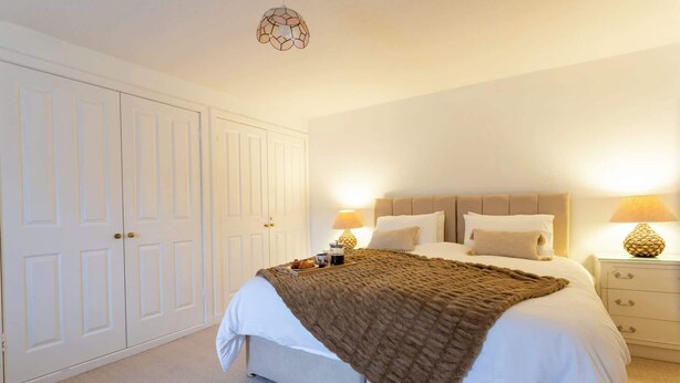 A bedroom with a large bed, two bedside tables with lamps, a wardrobe, and a ceiling light fixture. - New Cross Keys House - Cosy 4-bed Cottage in Kirk Yetholm (Kelso)