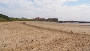 Una playa arenosa con barcos, un edificio grande y una duna al fondo.