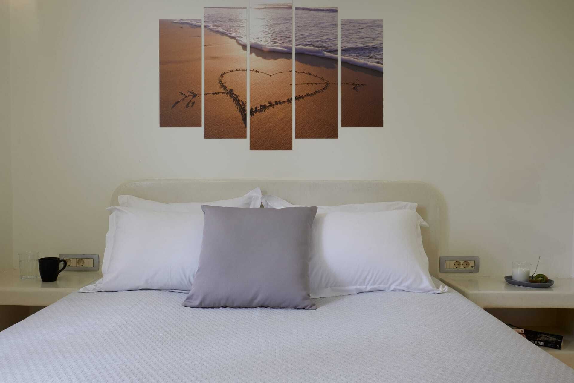 A bedroom with a bed, white bedding, a grey pillow, a wall-mounted artwork of a beach scene, and a bedside table with a black mug and a small plant.