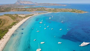 An aerial view of a coastal area with clear turquoise waters, numerous boats, and a prominent mountain in the background. - Apposentu (Case Peschiera-Lu Fraili)