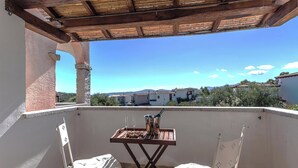 A balcony with a wooden table, two white chairs, and a view of buildings and mountains in the background. - Apposentu (Case Peschiera-Lu Fraili)