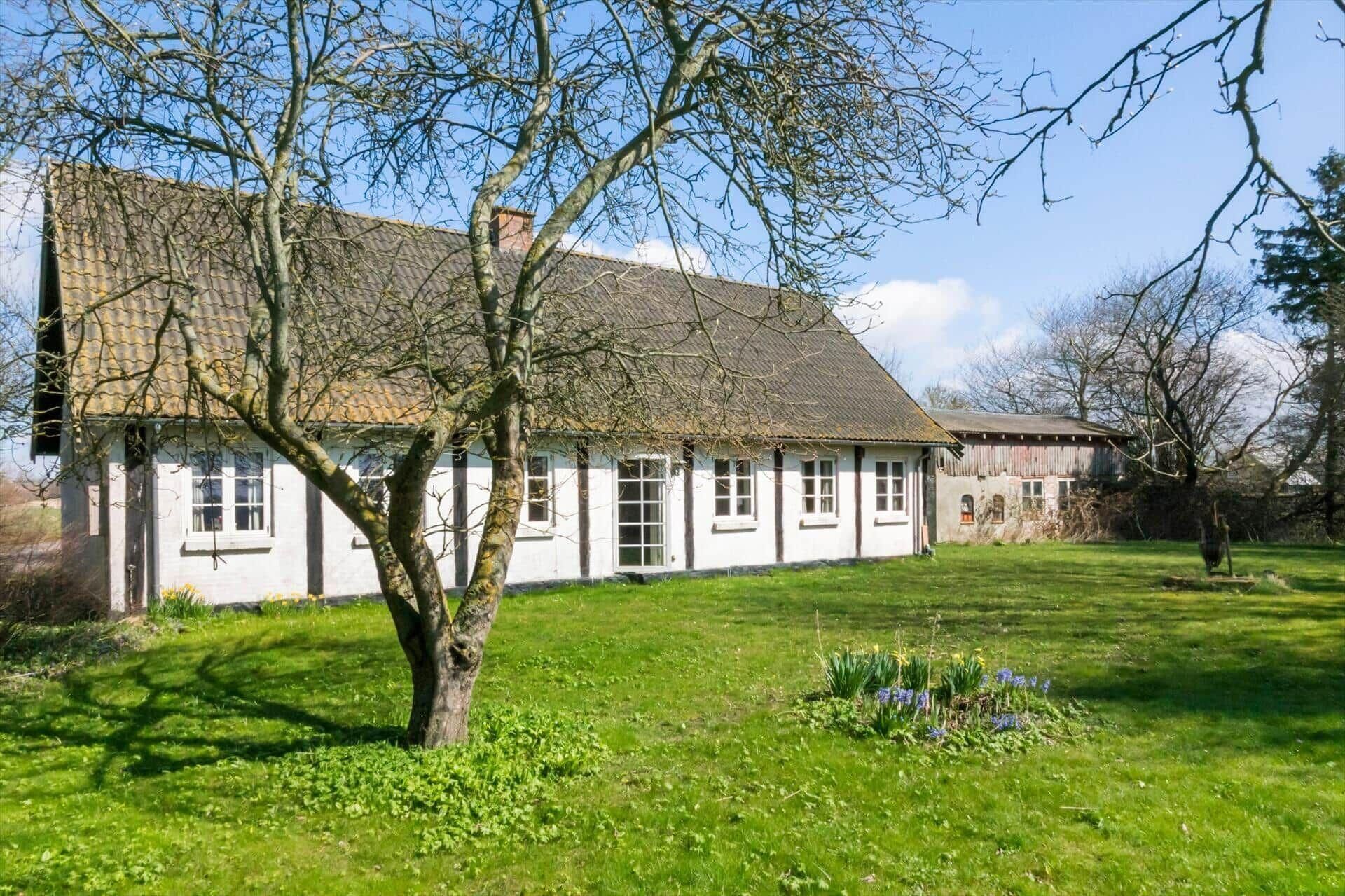 Un bâtiment traditionnel de couleur blanche, doté d’un toit en pente, entouré d’une zone herbeuse avec un arbre et des fleurs.