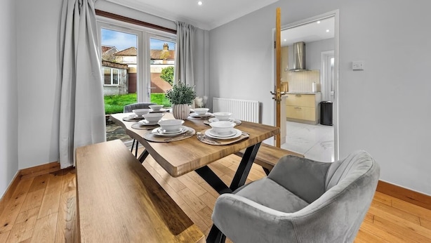 A dining area with a wooden table set for four, a grey upholstered chair, and a view of a garden through a window. - Modern 3BR London Home Pet Friendly with Private Garden Free Parking and WiFi (London)