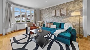A modern living room with a teal sofa, a coffee table, and a view of houses through large windows. - Modern 3BR London Home Pet Friendly with Private Garden Free Parking and WiFi (London)