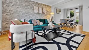 A modern living room with a brick wall, a sofa, a chair, a coffee table, and a dining area with a table and chairs. - Modern 3BR London Home Pet Friendly with Private Garden Free Parking and WiFi (London)