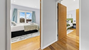 A bedroom with a bed, a window with curtains, a wooden floor, and a view of another room. - Modern 3BR London Home Pet Friendly with Private Garden Free Parking and WiFi (London)