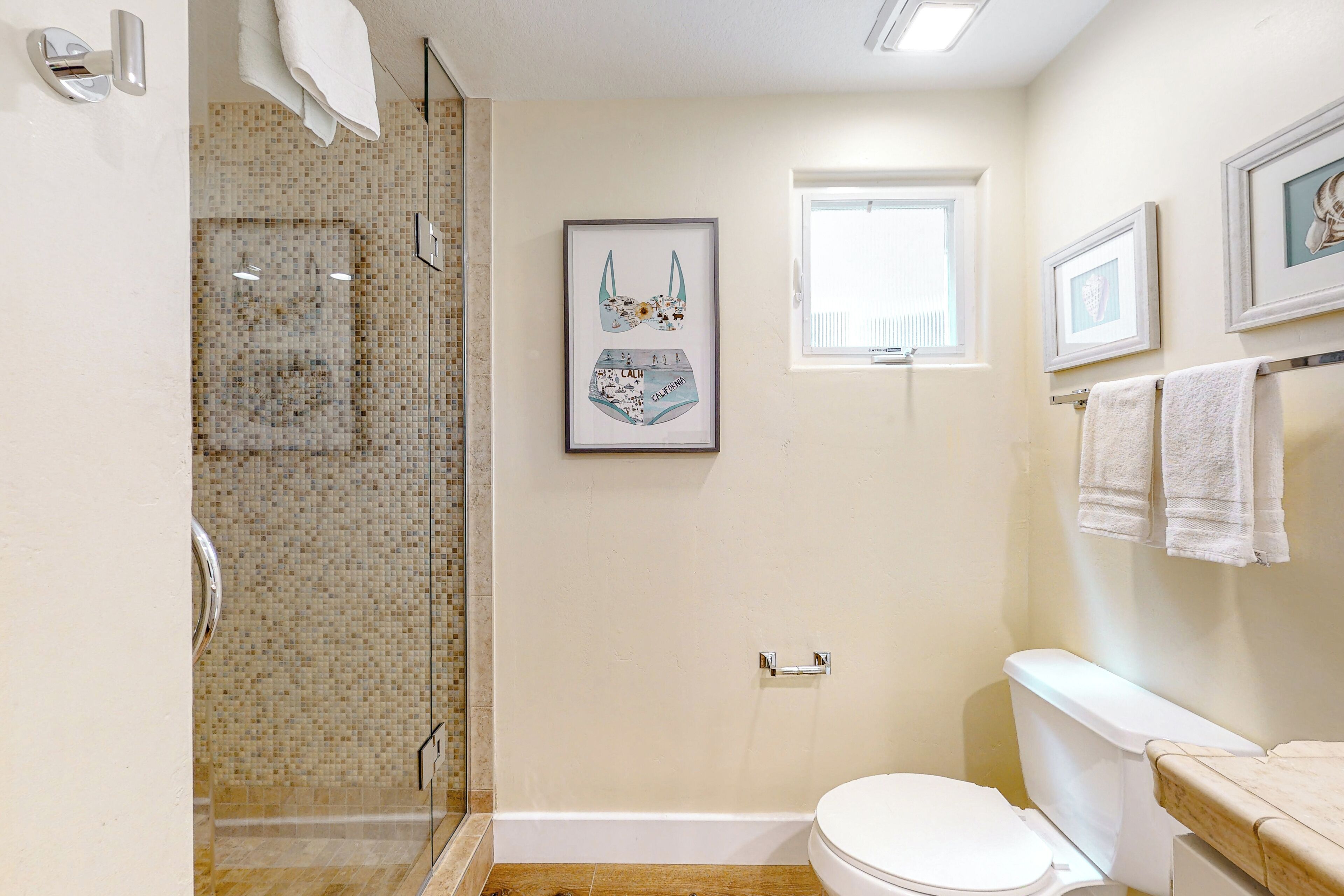 A bathroom with a glass shower, a toilet, and a sink. There are framed pictures on the wall and a window.