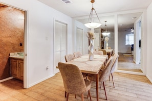 Uma sala de jantar com mesa e cadeiras de madeira, luminária pendente e cozinha visível por uma porta aberta.
