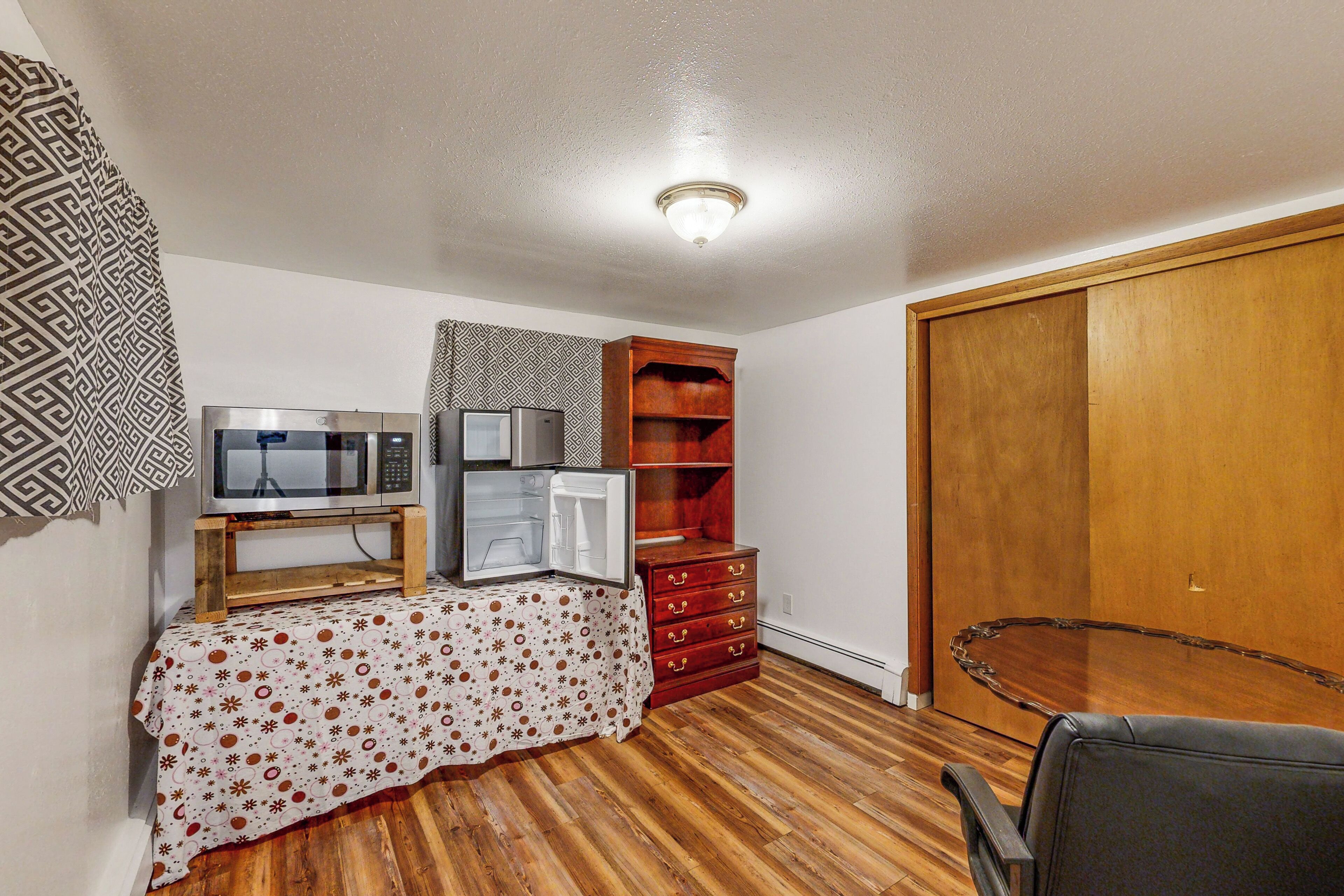 A small room with wooden flooring, a microwave, a refrigerator, a wooden cabinet, and a black chair.