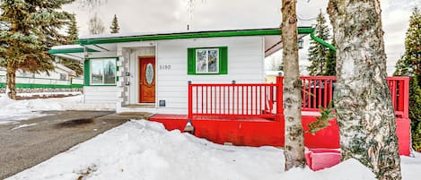 A white building with green trim and a red porch, surrounded by snow and trees.