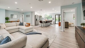Un vaste espace de vie comprenant un canapé d’angle, une table à manger avec des chaises, une cuisine équipée d’appareils électroménagers modernes et une vue sur l’extérieur.