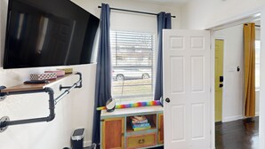 A room with a TV mounted on the wall, a window with curtains, a door, and a small wooden cabinet with storage boxes. - 2 BR Home one Block From Botanical Gardens (San Antonio)
