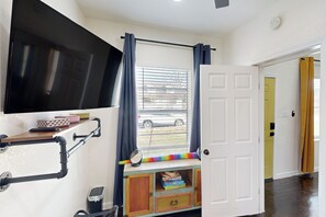 Ein Zimmer mit an der Wand befestigtem Fernseher, einem Fenster mit Vorhängen, einer Tür und einer kleinen Holzkommode mit Aufbewahrungsboxen.