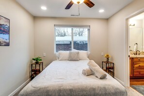 A bedroom with a large bed, a ceiling fan, a window with a view of a brick wall and trees, and a bathroom with wooden cabinets. - Denver Home w/ Patio Near City Park & Cherry Creek (Denver)