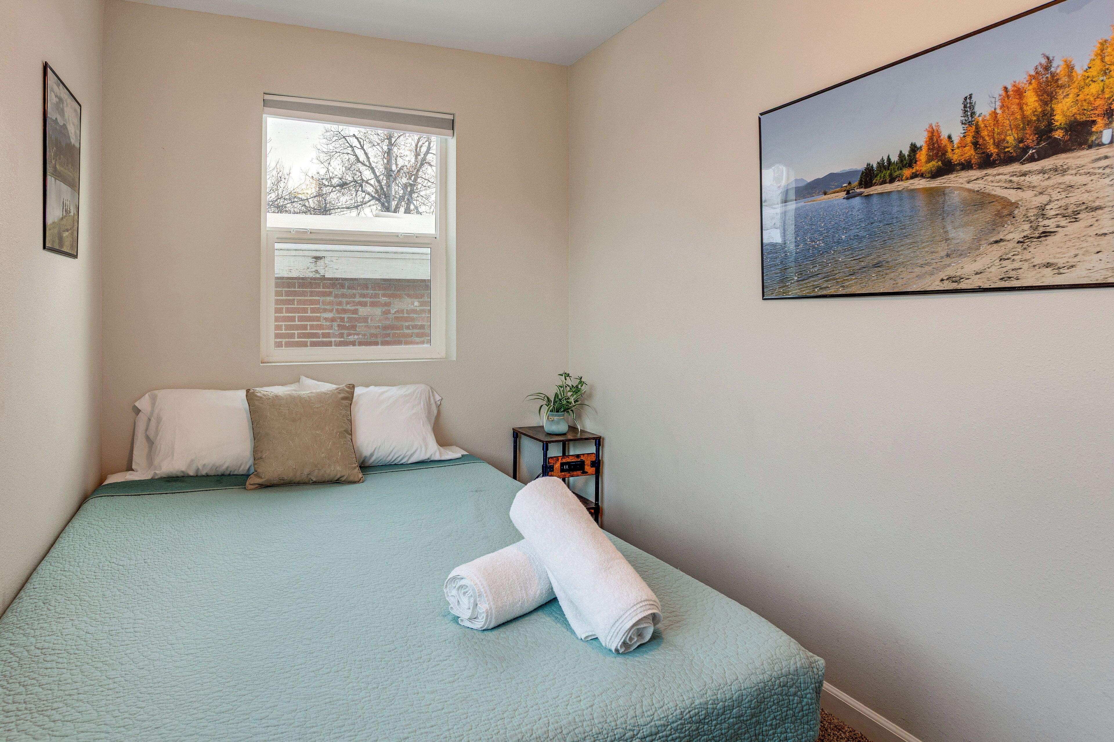 A bedroom with a bed, pillows, a window, a plant, and a painting.