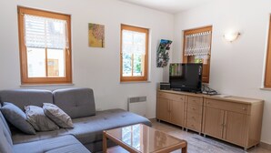 A living room with a grey sofa, a wooden coffee table, a TV on a wooden cabinet, and a window with lace curtains. - House Parabola – 4-Bedroom Mountain Retreat with Breathtaking Views, Sleeps 10 (Log pod Mangartom)