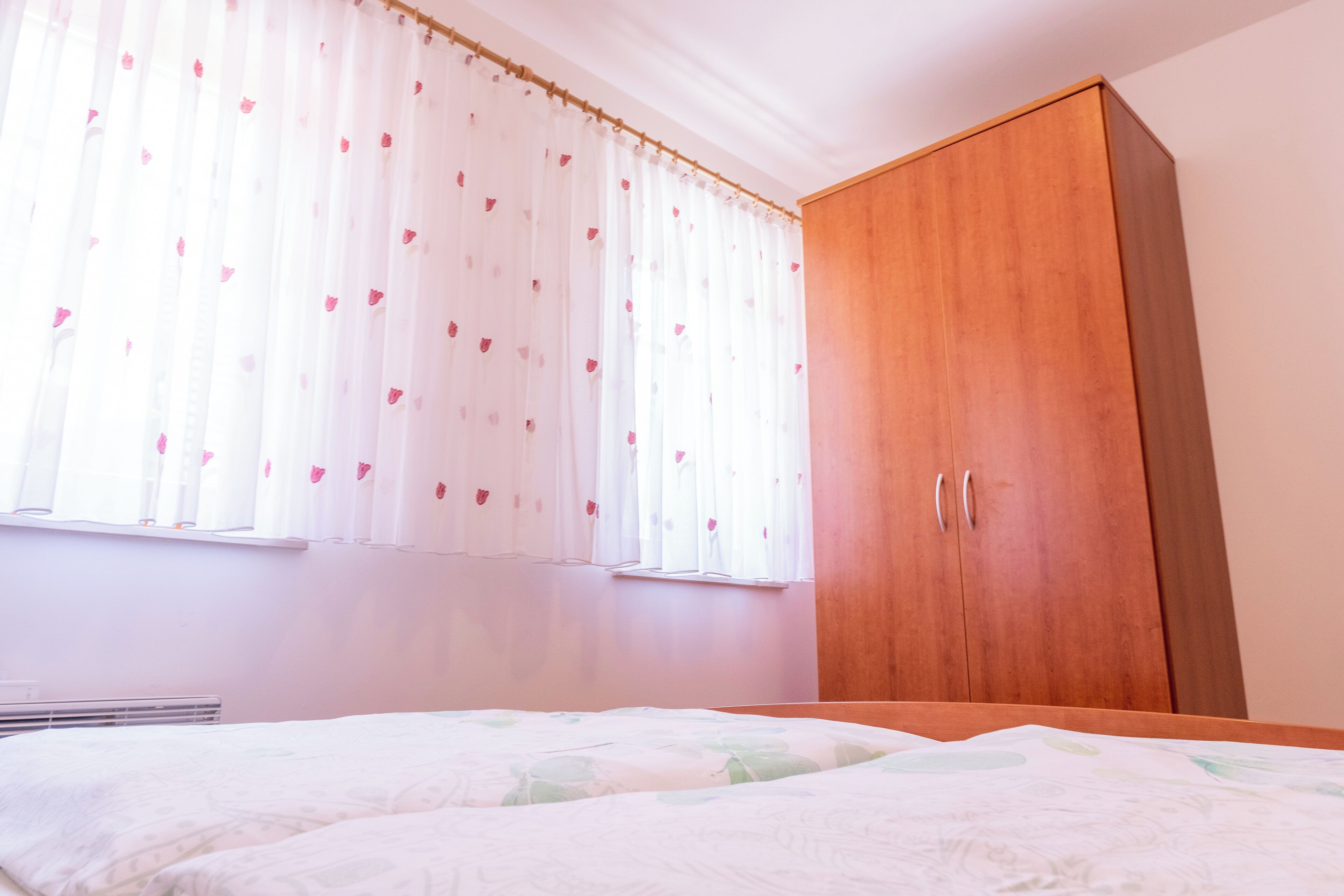 A bedroom with a wooden wardrobe, a bed with a floral bedspread, and sheer curtains with a heart pattern.