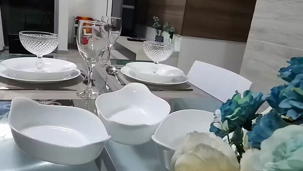 A modern dining area with a long table set for a meal, featuring white plates, bowls, and glasses.
